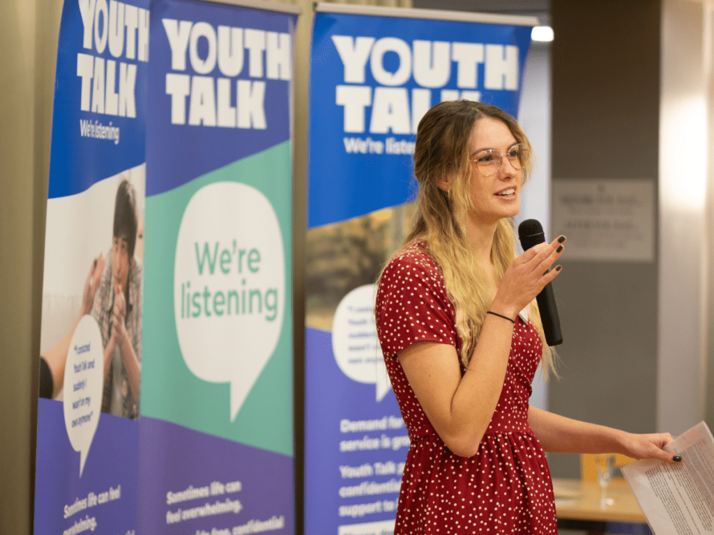 Local young person Rosie holding a microphone sharing her experience with mental health in front of Youth Talk banners at our 25th anniversary event