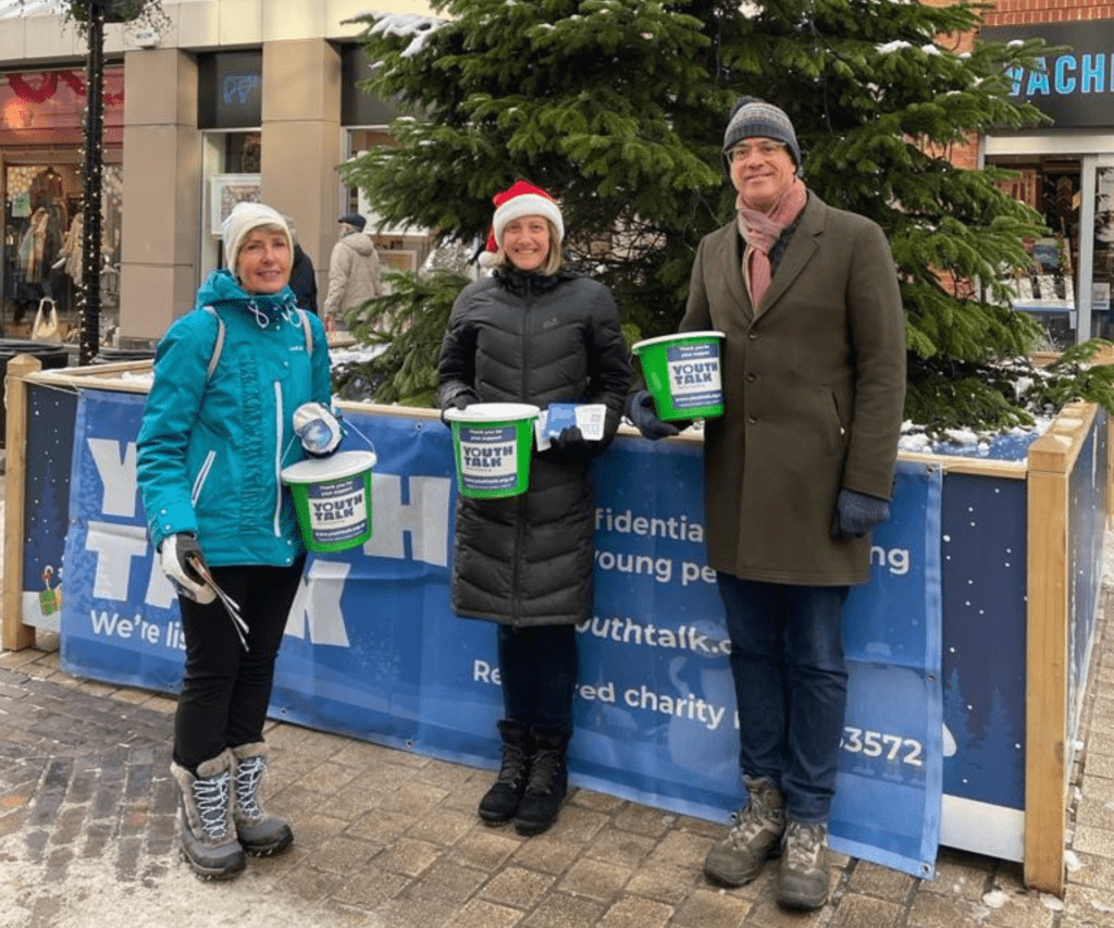 Our Corporate and Community Fundraiser, Jacqueline, standing in front of a Christmas tree holding collection buckets with some Youth Talk volunteers.