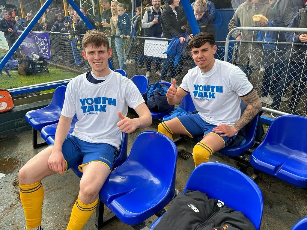 Two St Albans City football players sitting on chairs wearing Youth Talk t-shirts.