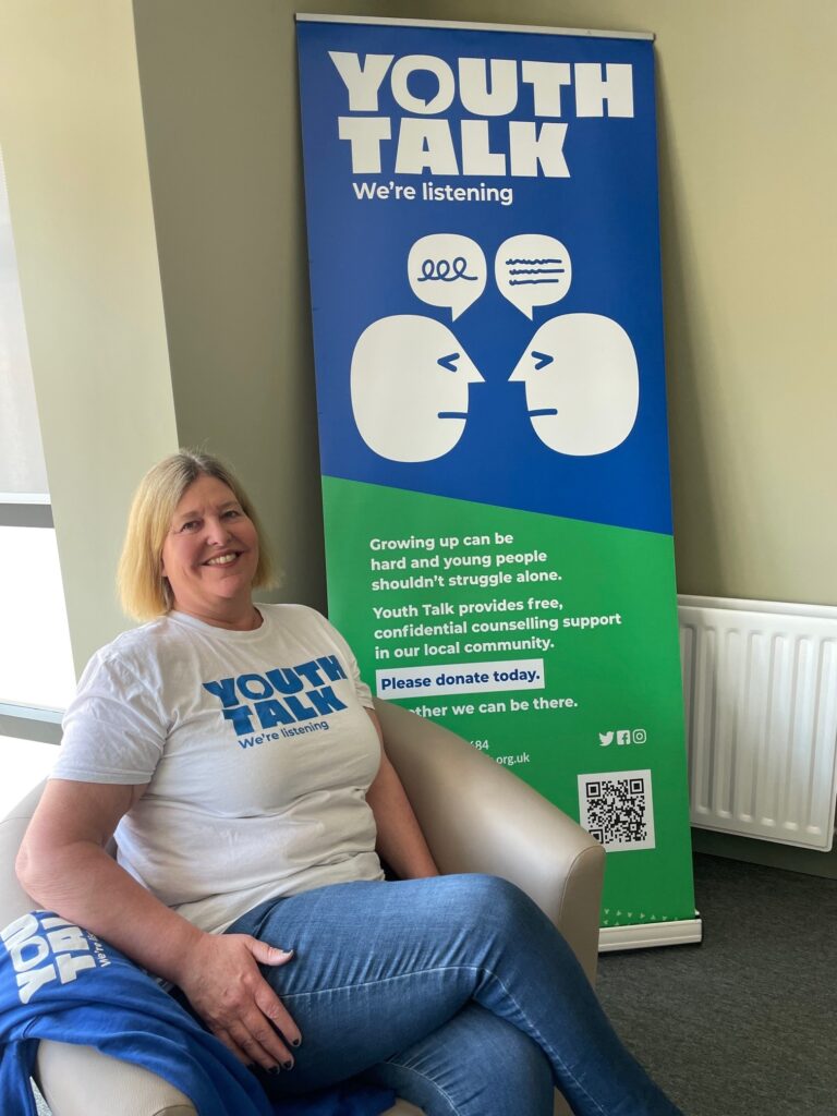 Our Volunteer Coordinator, Julia, sitting in front of a Youth Talk pop-up banner and wearing a white t-shirt with Youth Talk's logo on it.
